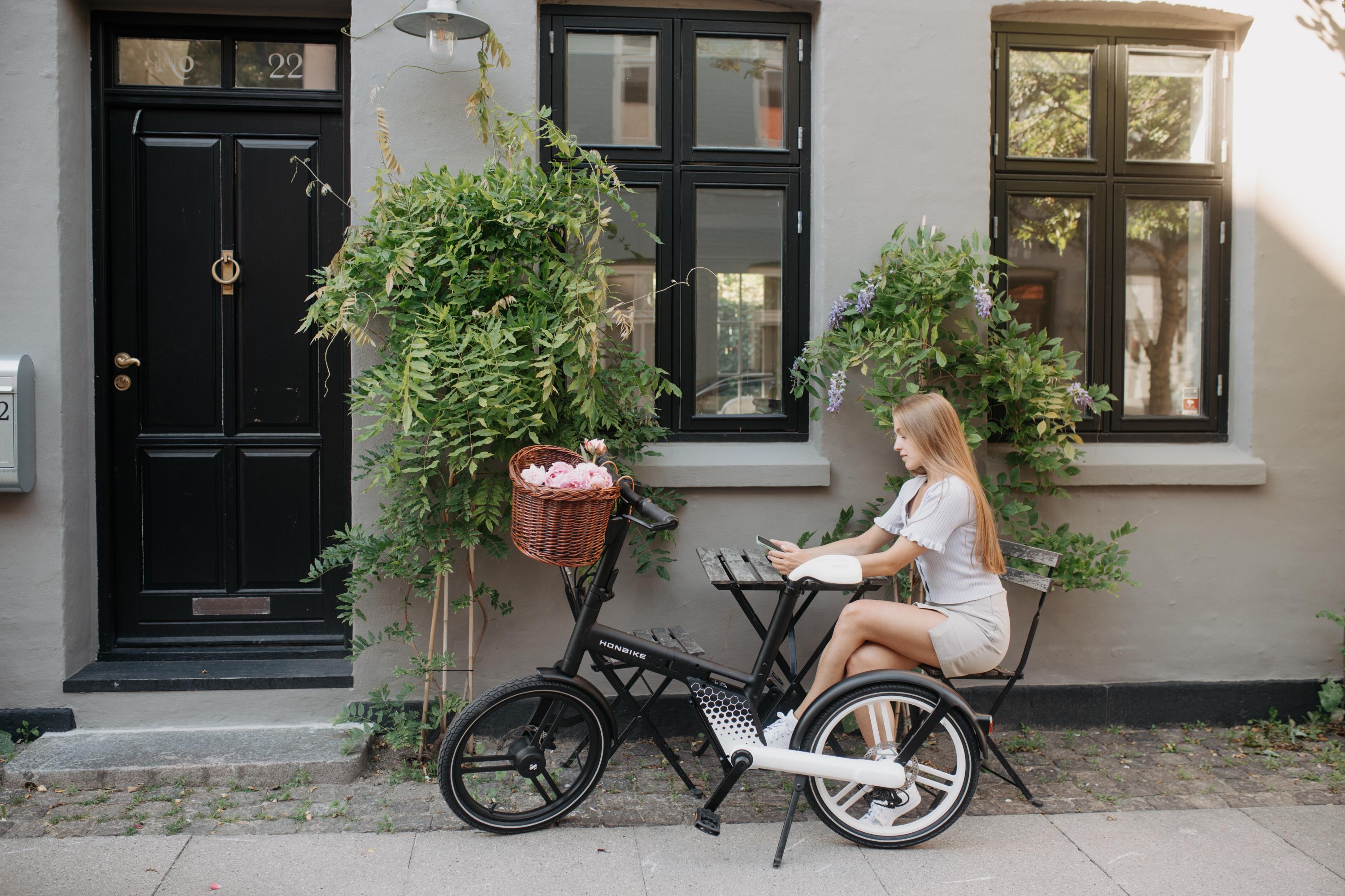 白い建物の前で、女性が電動自転車に乗り、スマートフォンを持っている。籠には花が入っている。
