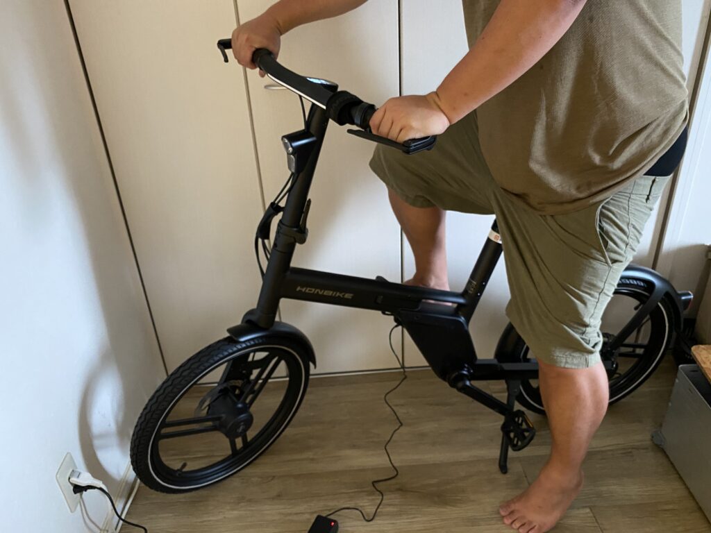 男性がHONBIKEというブランドの黒い電動折りたたみ自転車を持ち、室内で充電している様子。