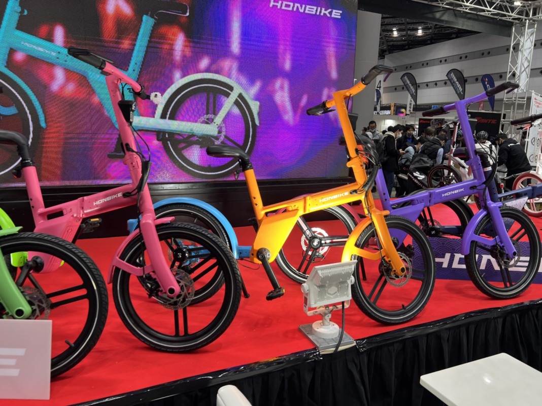 HONBIKE のカラフルな電動バイクが展示されているショー会場。複数の鮮やかな色のモデルが赤いステージに並んでいる。